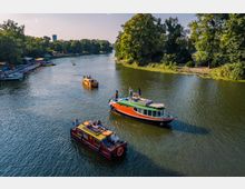 Flusslandschaft mit kleinen Freizeitbooten, umgeben von dichtem Baumbestand auf beiden Seiten. Am Ufer links sind bunte Tretboote vertäut, während mehrere Boote mit Menschen auf dem Wasser unterwegs sind.
