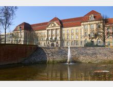Historisches Verwaltungsgebäude mit beige-gelber Fassade im neoklassizistischen Stil, rotem Satteldach und verziertem Eingang, im Vordergrund ein Wassergraben mit einer kleinen Springbrunnenfontäne.