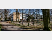 Ein kleiner Spielplatz mit Klettergerüst und einer Wippe, umgeben von einem grünen Metallzaun und Bänken, steht in einem parkähnlichen Bereich mit Bäumen. Im Hintergrund ist ein modernes, mehrstöckiges Gebäude mit gelber Fassade und blauen Dächern zu sehen, das teilweise mit Graffiti besprüht ist.