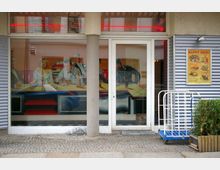 Eine Fensterfront eines Restaurants oder Cafés mit einem gemalten Wandbild, das berühmte Persönlichkeiten an einem Tisch zeigt, und einem angrenzenden Werbeplakat für Speisenangebote. Vor dem Eingang stehen ein blauer Transportwagen und ein Pflanzkübel.