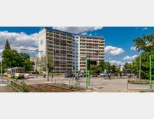 Mehrstöckiges Wohngebäude mit Balkonreihen im Vordergrund, daneben eine Straßenbahn-Haltestelle und Gleise. Im Umfeld befinden sich eine Kreuzung, einige Bäume und einzelne Passanten bei sonnigem Wetter.