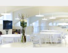 Heller, moderner Veranstaltungsraum mit runden Tischen, weißen Tischdecken und Stühlen. Im Vordergrund steht eine Glasvase mit weißen Blumen und Kätzchenzweigen; im Hintergrund sind geschwungene Deckenelemente und große Fenster zu sehen.
