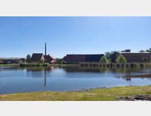 Eine historische Backsteinanlage mit mehreren Gebäuden, darunter ein Gebäude mit Schornstein, spiegelt sich in einem ruhig gelegenen See. Im Vordergrund ist eine grasbewachsene Uferlandschaft sichtbar, im Hintergrund Bäume und ein klarer blauer Himmel.