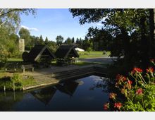 Grüne Parklandschaft mit einem kleinen Teich im Vordergrund, umgeben von Pflanzen und roten Geranien. Im Hintergrund befinden sich mehrere überdachte Sitzbereiche mit Holzdächern, Bänken und Tischen sowie offene Grasflächen und Bäume.