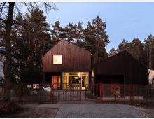 Moderne Wohngebäude mit dunkler Holzfassade, bestehend aus zwei Giebelhäusern nebeneinander. Im Vordergrund befindet sich ein Metallzaun mit Tor, dahinter eine beleuchtete Glasfront des Haupthauses; umgeben von hohen Kiefern.