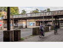 Eine Straßenunterführung mit einer alten Eisenbahnbrücke, deren Metallträger mit Graffiti und Schriftzügen bemalt sind. Im Vordergrund fahren zwei Personen auf Fahrrädern auf einem gepflasterten Gehweg, während im Hintergrund teils bewachsene Backsteinwände mit Plakaten und Grafitti sichtbar sind.