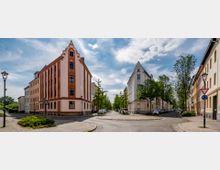 Straßenkreuzung in einer historischen Wohngegend mit mehrstöckigen Gebäuden im Stil der Gründerzeit. Die Fassaden sind verziert und pastellfarben, umgeben von gepflasterten Gehwegen, Bäumen und geparkten Autos.