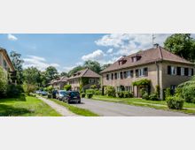 Straßenszene in einem Wohngebiet mit zweistöckigen Mehrfamilienhäusern aus der Nachkriegszeit, charakterisiert durch rote Ziegeldächer, helle Fassaden und Fensterläden. Die Straße wird von parkenden Autos gesäumt, während Gärten und Bäume das Viertel umgeben.