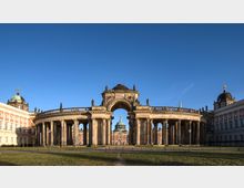 Historisches Gebäudeensemble mit einer halbkreisförmigen Kolonnade, in der Mitte ein monumentales Tor mit Kuppel, flankiert von Gebäuden mit verzierten Fassaden und Kuppeln. Der Vordergrund zeigt eine Grünfläche unter einem klaren blauen Himmel.