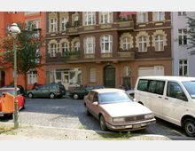 Straßenszene mit gepflastertem Platz, geparkten Autos und einem mehrstöckigen Wohnhaus im Stil der Gründerzeit mit verzierten Fenstern und Balkonen. Im Vordergrund steht eine alte Laterne, und im Hintergrund grenzen weitere bunte Gebäude an.