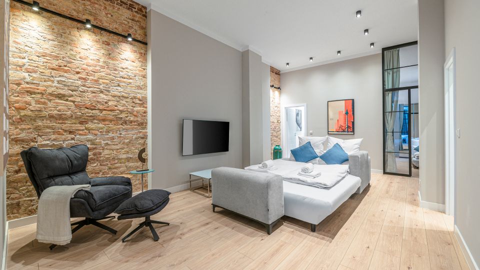 Modernes, helles Schlafzimmer mit freiliegender Backsteinwand, hohen Decken und hellem Holzboden. In der Mitte steht ein graues Polsterbett mit Kissen; daneben sind ein schwarzer Fernseher an der Wand, ein Sessel mit Hocker und eine Glas-Metall-Schiebetür zu einem Nebenraum.