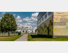 Gebäude mit gelb-grüner Fassade rechts im Vordergrund, ein gepflasterter Weg führt durch eine Rasenfläche entlang eines Baums zu einem eingezäunten Sportplatz; im Hintergrund sind moderne Wohn- und Bürogebäude zu sehen vor einem bewölkten Himmel.