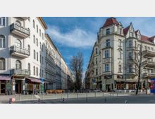 Straßenszene mit Altbauten, die beidseitig die Straße säumen, mit verzierten Fassaden und Balkonen. Im Vordergrund befinden sich Cafés und Geschäfte, während im Hintergrund ein kahler Baum in einer verkehrsberuhigten Zone steht.