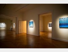 Innenraum einer modernen Kunstgalerie mit Holzboden und cremefarbenen Wänden. Mehrere farbige Gemälde hängen an den Wänden, und die Räume sind durch offene Türen miteinander verbunden, beleuchtet von gerichteten Deckenstrahlern.