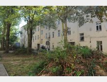 Mehrstöckiges, verblasstes Wohngebäude mit sichtbaren Alters- und Gebrauchsspuren, umgeben von hohen, dicht belaubten Bäumen. Im Vordergrund befinden sich überwucherte Grünflächen und abgeschnittenes Astwerk.