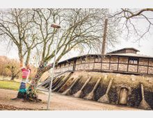 Ein historisches Gebäude mit Fachwerk und Backsteinfassade, flankiert von einem mit bunten, gestrickten Stoffelementen dekorierten Baum. Im Vordergrund ein befestigter Weg und eine Laterne, umgeben von kahlen Bäumen in einer Parklandschaft.