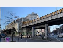 Straßenszene mit einer erhöhten U-Bahn-Station in einem historischen Baustil, erkennbar an den großen Glasfenstern und den Verzierungen. Eine gelbe U-Bahn fährt über die Brücke, während Fußgänger und Autos die Straße darunter benutzen.