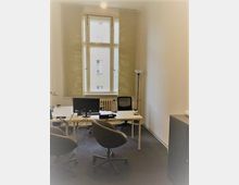 Büro mit zwei Schreibtischen, auf denen ein Computerbildschirm und Arbeitsmaterialien stehen. Große Fenster mit halb heruntergelassenen Rollos lassen Tageslicht herein, und im Raum stehen Bürostühle auf einem Teppichboden.