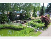 Ein grüner Garten mit einem kleinen Teich, der von Schilf umgeben ist, einer kleinen Brücke aus Metall und Sitzmöbeln aus Holz und Rattan. Im Hintergrund stehen Bäume und eine Überdachung sowie ein Bauzaun neben parkenden Autos.