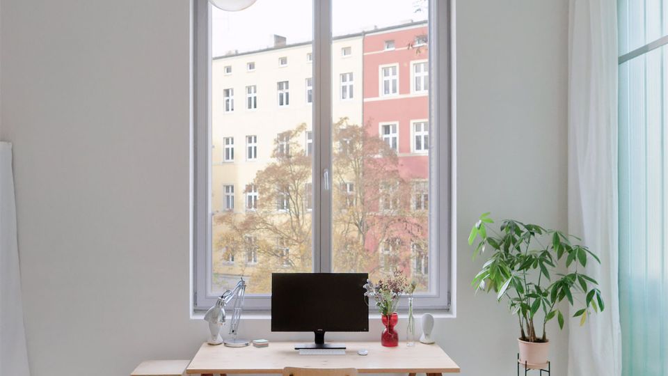 Helles, minimalistisches Homeoffice mit großem Fenster zur Straße, schlichtem Schreibtisch auf Holztresteln, Monitor und Lampe sowie einem einzelnen Stuhl. Rechts stehen eine hohe Zimmerpflanze und bodenlange weiße Vorhänge neben einer türkisfarbenen Glasfront.