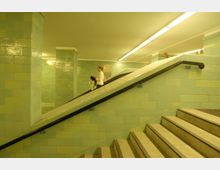 Unterführung mit hellgrün gefliesten Wänden und Treppen, die auf beiden Seiten durch schwarze Handläufe begrenzt sind. Im Hintergrund befindet sich eine Rampe, auf der Menschen gehen, unter einer weißen Deckenbeleuchtung.