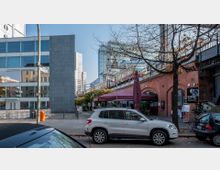 Straßenszene mit modernen und historischen Gebäuden: Links steht ein Bürogebäude mit großen Glasfassaden, während rechts ein rotes Klinkergebäude einen Bahndamm trägt. Eine Bahn fährt über den Damm, ein rotes Sonnensegel und mehrere parkende Autos prägen den Vordergrund.