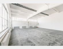 Leerer, heller Raum mit glatten, grauen Betonböden und weißen Wänden. Große Fenster und Deckenstreben aus schwarzem Metall prägen die Industrie-Ästhetik der Halle.