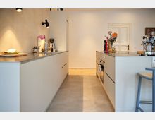 Moderne Küche mit hellen, glatten Einbaufronten, zwei gegenüberliegenden Arbeitsflächen und einer Kücheninsel mit Kochfeld. Im Hintergrund befindet sich eine weiße Tür, und auf den Arbeitsflächen stehen Geräte, Deko-Objekte sowie eine Kaffeemaschine.