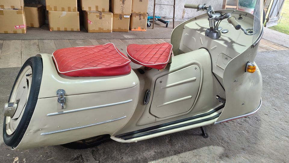 Innenraum einer Lager- oder Werkstatthalle mit Betonboden und Holzplanken, im Hintergrund stapelweise Kartons sowie eine Leiter. Im Vordergrund steht ein helles Vintage-Motorroller-Gespann mit roter Sitzpolsterung und kleiner Windschutzscheibe.