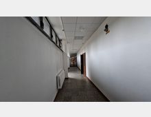 Langer, schmaler Innenflur in einem Gebäude mit weißen Wänden, grauem Steinboden und abgehängter Rasterdecke mit Leuchten. Links befinden sich hohe Fenster und ein Heizkörper, rechts mehrere Türen; am Ende führt eine verglaste Tür in einen weiteren Bereich.