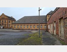 Innenhof eines alten Backstein-Industriekomplexes mit langem, niedrigem Gebäude und Satteldach sowie einem mehrstöckigen Ziegelbau im Hintergrund. Im Vordergrund liegt eine große asphaltierte Fläche mit Moosstellen und einem hohen Mast; rechts ist eine teilweise beschädigte Ziegelwand mit zugemauerten Öffnungen zu sehen.