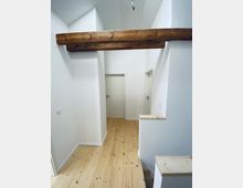 Flur mit hellen Holzfußbodenbrettern, weiß gestrichenen Wänden und einer freiliegenden, rustikalen Holzbalkendecke. Der Raum führt zu mehreren weißen Türen.