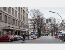 Straßenszene in einer Stadt mit mehrstöckigen Gebäuden im Hintergrund, einem breiten Gehweg mit kahlen Bäumen und einem angrenzenden historischen Gebäude aus Stein. Im Vordergrund fahren Autos, ein Fahrradfahrer ist unterwegs, und Straßenschilder weisen auf einen kombinierten Geh- und Radweg hin.