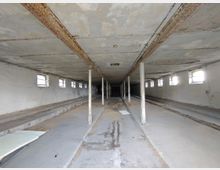 Verlassener, rechteckiger Innenraum mit niedriger Decke und rostigen Metallträgern, unterstützt von Metallstützen. Der Raum hat kleine, rechteckige Fenster an den Wänden, einen Betonboden mit Gräben in der Mitte und sichtbare Verfallsspuren.