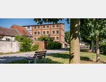 Blick auf einen kleinen Grünstreifen mit Parkbank und Bäumen neben einem Fußweg, der zu einem mehrstöckigen Backstein-Industriegebäude mit vielen Fenstern führt. Links stehen Fachwerk- und Ziegelhäuser, entlang des Weges sind Büsche und eine Straßenlaterne zu sehen.