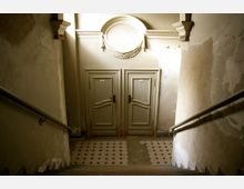 Treppenhaus mit abgenutzten Wänden und einem Fliesenboden in geometrischem Muster. Am Ende der Treppe befinden sich zwei grüne Holztüren mit geschwungenen Details und ovalem Dekorelement darüber.