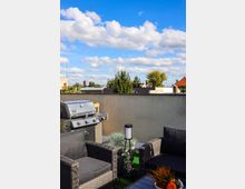 Eine Dachterrasse mit modernen Gartenmöbeln aus dunklem Geflecht, einem Grill aus Edelstahl und dekorativen Pflanzen in Töpfen, umgeben von einer niedrigen Mauer. Im Hintergrund sind Bäume, Wohngebäude und ein blauer Himmel mit weißen Wolken sichtbar.