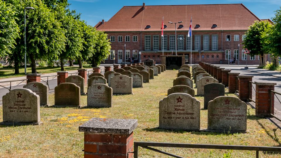 Reihen von Grabsteinen auf einer rechteckigen, von niedrigen Ziegelpfeilern und Metallgeländern eingefassten Grünfläche, flankiert von Bäumen. Im Hintergrund steht ein großes Backsteingebäude mit rotem Ziegeldach, großen Fenstern und Flaggenmasten.