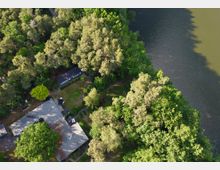 Luftaufnahme eines Hauses mit Flachdach, umgeben von dichter Waldlandschaft und einer angrenzenden Wasserfläche. Der Garten des Hauses ist teilweise erkennbar, mit Rasen, Bäumen und einer Terrasse mit Möbeln.