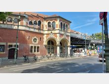 Rotes Backsteingebäude mit historischer Architektur, verziert durch Rundbögen, Säulen und kleine Fenster unterhalb eines Steildachs, daneben eine erhöhte U-Bahn-Brücke mit einem Schild der Station "Schlesisches Tor". Fahrräder sind vor dem Gebäude abgestellt, während weitere Straßenelemente, wie Laternen und Verkehrsschilder, die Umgebung prägen.