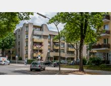 Straßenszene in einem Wohngebiet mit mehrstöckigen Wohngebäuden, die schlichte beige Fassaden und Balkone mit Blumendekoration aufweisen. Im Vordergrund stehen parkende Autos, Bäume spenden Schatten, und Verkehrsschilder markieren eine Tempo-30-Zone.