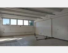 Leerer Raum mit weißen Fliesen an den Wänden und dem Boden, großen Fenstern an einer Seite, einer darunterliegenden Heizkörperverkleidung und sichtbaren Rohren sowie Leitungen nahe dem Boden. Die Decke ist mit freiliegenden Balken und langen Hängeleuchten ausgestattet.