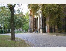 Klassizistischer Kircheneingang mit hohen dunklen Säulen und einer großen zweiflügeligen Holztür. Im Vordergrund gepflasterter Weg, umgeben von Bäumen und Grünflächen, mit Fahrradständern und einer Person vor dem Eingang.