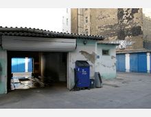 Ein kleiner Autowaschplatz mit einer überdachten Waschstation und einem blauen SB-Staubsauger an der Außenwand. Im Hintergrund sind blaue Garagentore sowie eine Wand mit abgebröckelter Fassade und Graffiti sichtbar.