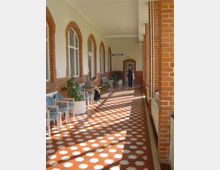 Ein langer, sonnendurchfluteter Flur mit cremefarbenen Wänden und roten Backsteinbögen um große Fenster. Der Boden ist mit rot-weißen, achteckigen Fliesen gestaltet, entlang der Wände stehen blaue Stühle und Topfpflanzen.
