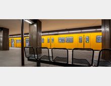 U-Bahn-Station mit gelbem U-Bahn-Wagen an einem Bahnsteig. Im Vordergrund stehen Metallbänke, während braune Säulen und eine helle Deckenbeleuchtung die Station strukturieren.