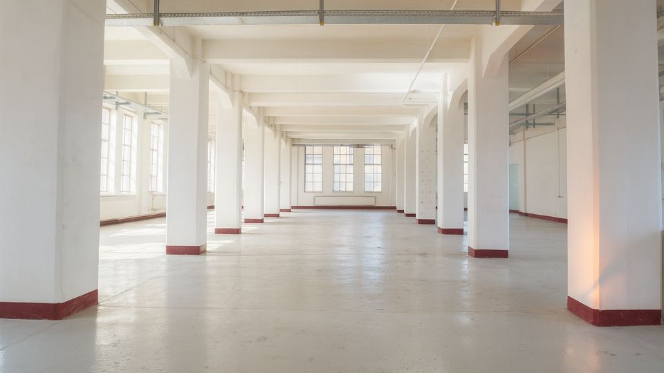 Großer, leerer Industriehallen- oder Loftinnenraum mit weiß gestrichenen Stützenreihen, sichtbaren Deckenbalken und Betonboden. Hohe Fenster entlang der Seiten lassen viel Tageslicht herein; rote Sockelleisten akzentuieren die Pfeiler.