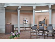 Innenraum einer schlichten Kirche mit hellen Wänden. Zu sehen sind eine Holzkanzel links, ein Ständer mit Liednummern in der Mitte, mehrere Stühle sowie eine kleine Orgel rechts im Hintergrund. Im Vordergrund steht ein Topf mit violetten Blumen.