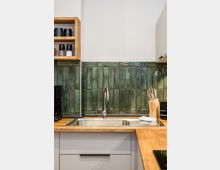 Moderne Küchenzeile mit Holzarbeitsplatte und Edelstahlspüle, darüber eine grün glasierte Fliesenrückwand. Links ein offenes Holzregal mit Gewürzstreuern, rechts ein Messerblock vor hellen Schränken.