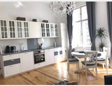 Helle Küche mit weißer Einbauzeile, Glastürenschränken, Holzarbeitsplatten und einem Edelstahl-Backofen. Rechts steht ein runder Esstisch mit vier Stühlen vor einem großen Fenster mit grauen Vorhängen; daneben befinden sich Zimmerpflanzen.